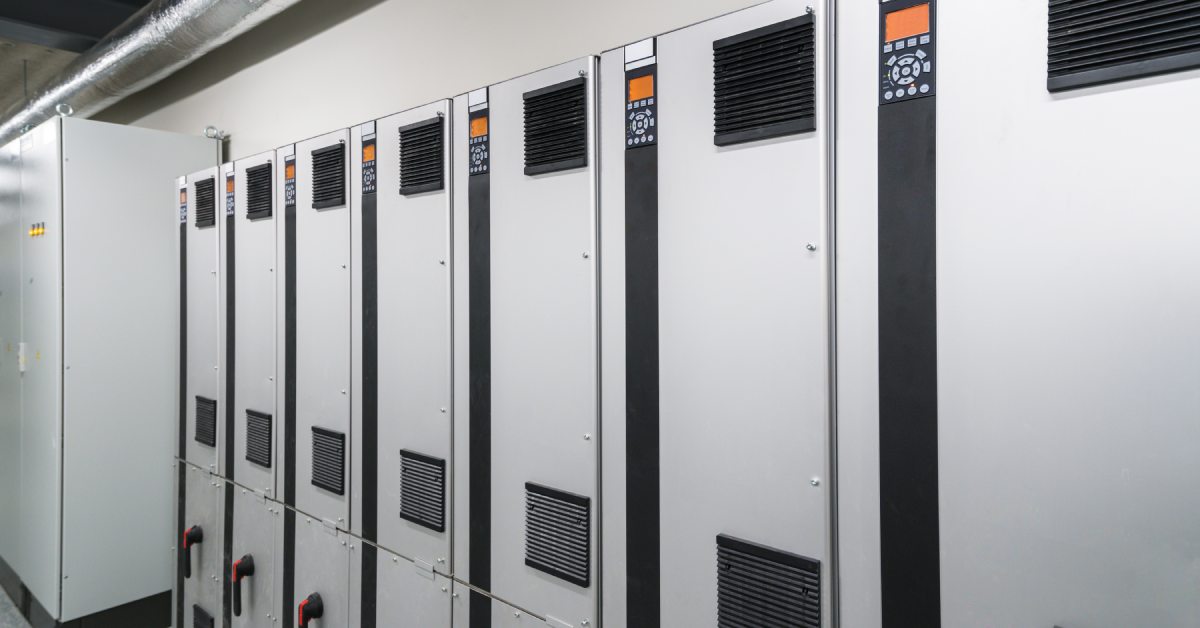 A row of seven connected silver variable speed drive inverters stands against a wall in an industrial setting.