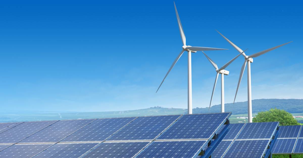 Three rows of solar panels are installed in front of three white wind turbines under a clear blue sky.
